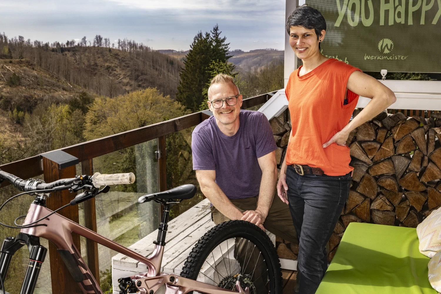 Zwei Personen mit Mountainbike auf einer Terrasse vor bewaldeten Hügeln