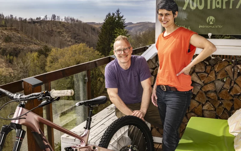 Zwei Personen mit Mountainbike auf einer Terrasse vor bewaldeten Hügeln